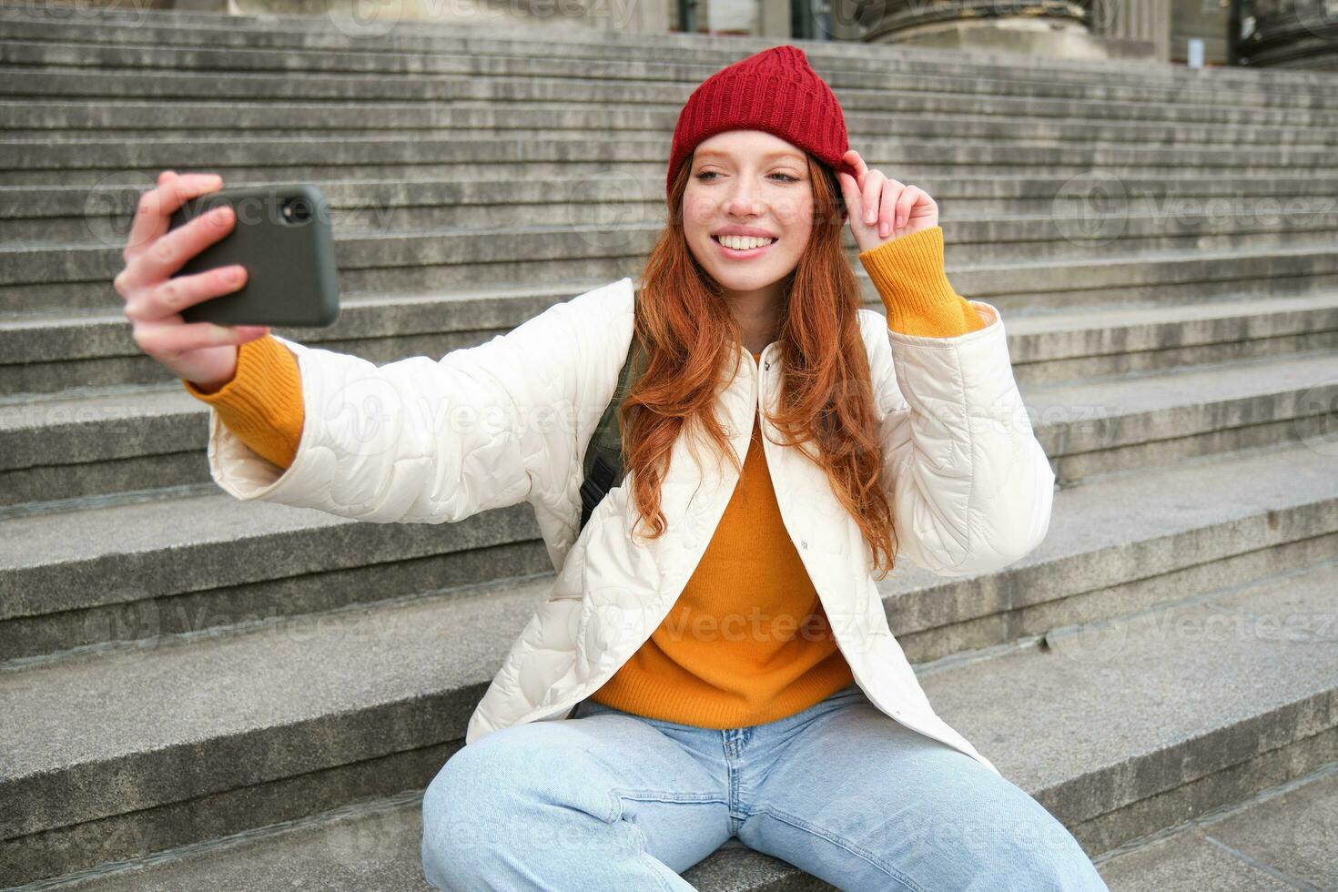 Stylish young girl in red hat, takes photos on smartphone camera, makes selfie as she sits on stairs near museum, posing for photo with app filter