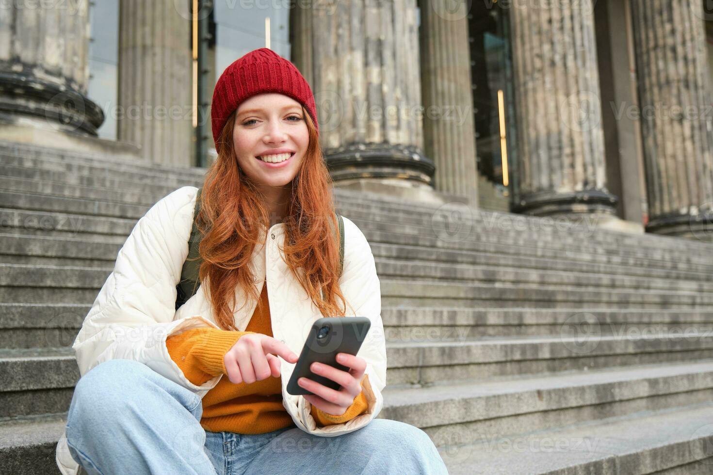 Portrait of young urban girl in red hat, sits on stairs near museum, holds mobile phone, connects to public wifi and surfs net, uses smartphone apps photo