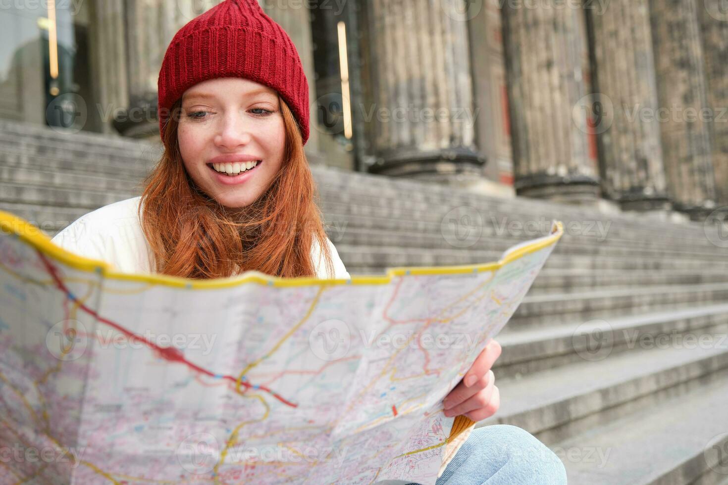 Smiling girl traveler, tourist sits on stairs near museum and holds paper map, explores city during sightseeing adventure trip photo