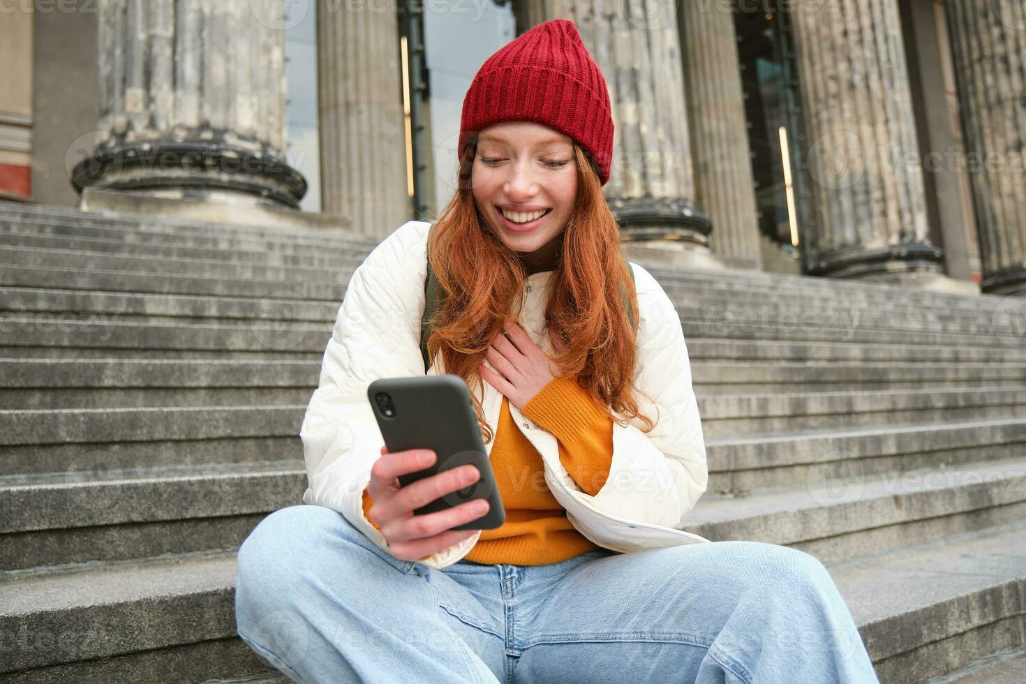 Young smiling woman with long red hair, sits near building on stairs with smartphone, using mobile application, sending message photo