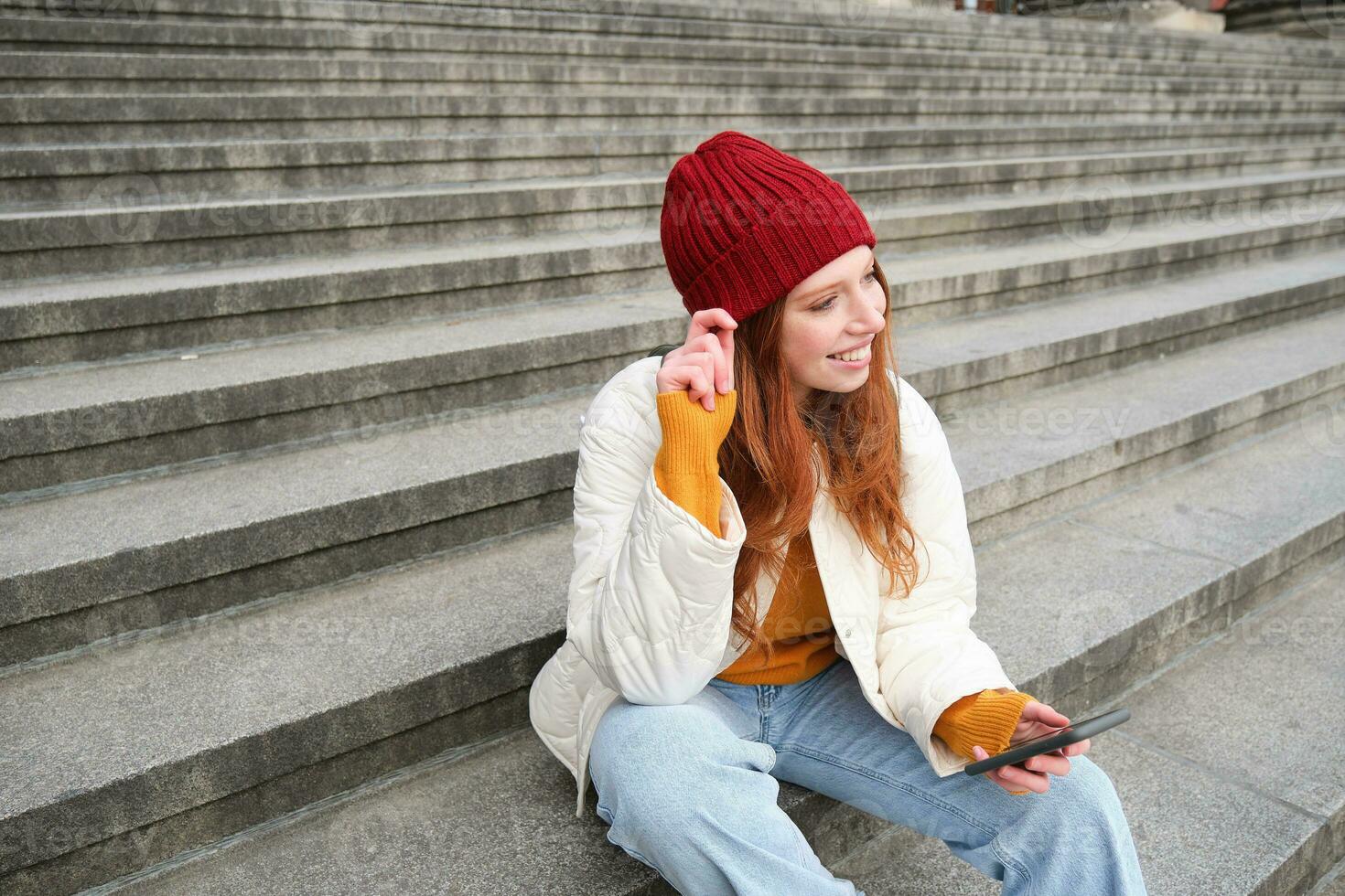 Portrait of young urban girl in red hat, sits on stairs near museum, holds mobile phone, connects to public wifi and surfs net, uses smartphone apps photo