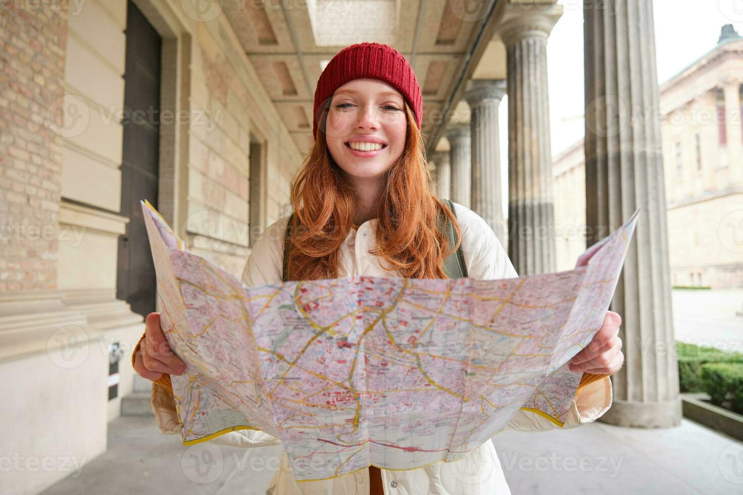 Adventurous redhead girl walks in town with paper map, explores city as tourist, looks for popular tourism attractions, looks around excited and smiles photo