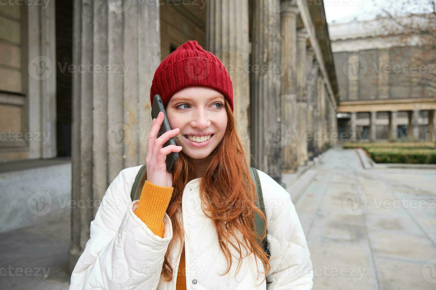 sonriente pelirrojo hembra turista negociaciones en móvil teléfono y camina alrededor ciudad. contento estudiante en rojo sombrero llamadas amigo, soportes en calle y usos teléfono inteligente foto