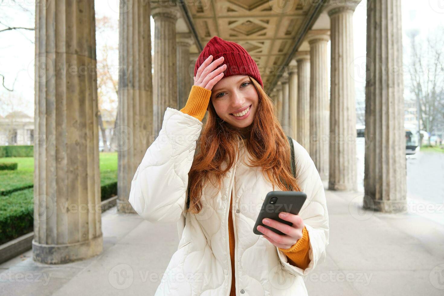 Happy young female model stands on street and holds mobile phone, uses smartphone app, follows the route of online map photo