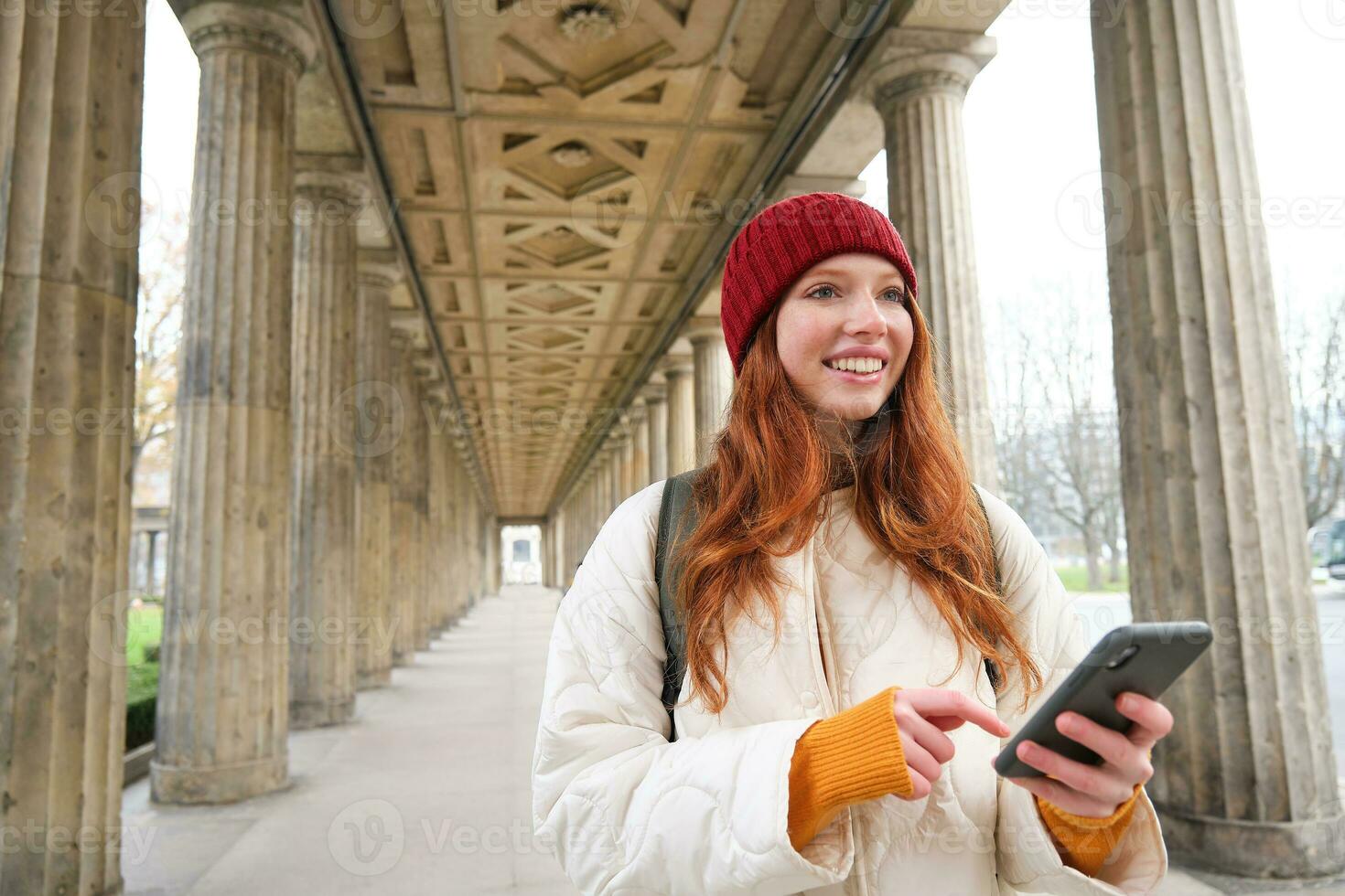 Happy young female model stands on street and holds mobile phone, uses smartphone app, follows the route of online map photo