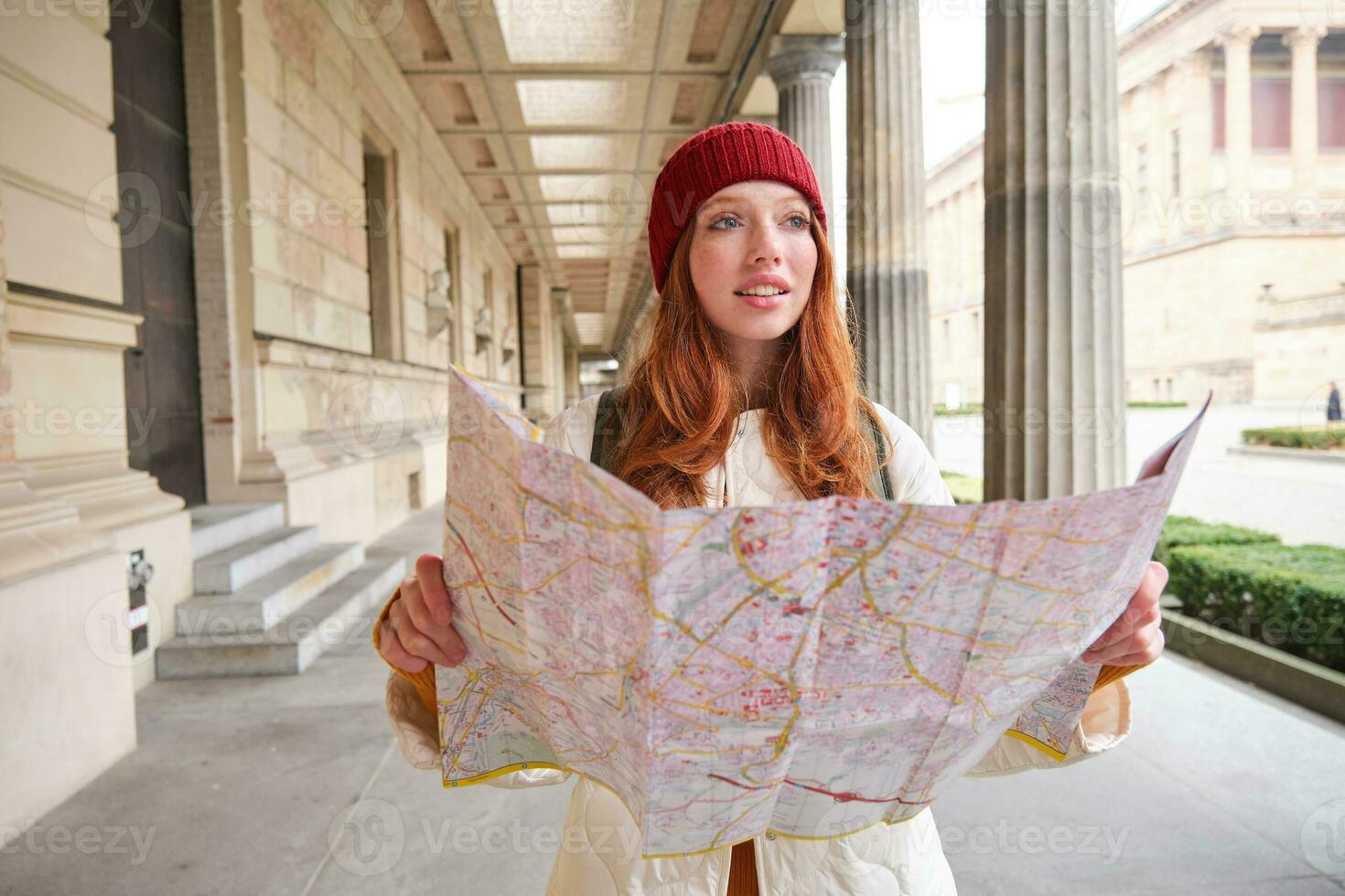 aventurero pelirrojo niña camina en pueblo con papel mapa, explora ciudad como turista, mira para popular turismo atracciones, mira alrededor emocionado y sonrisas foto