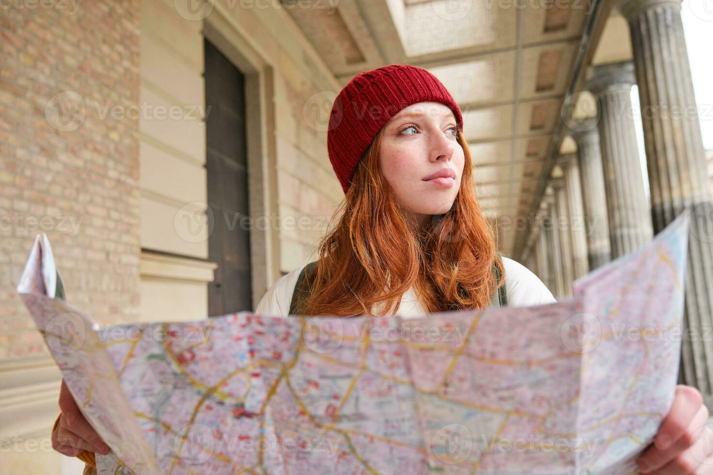 Smiling young redhead woman in red hat, looks at paper map to look for tourist attraction. Tourism and people concept. Girl explores city, tried to find way photo