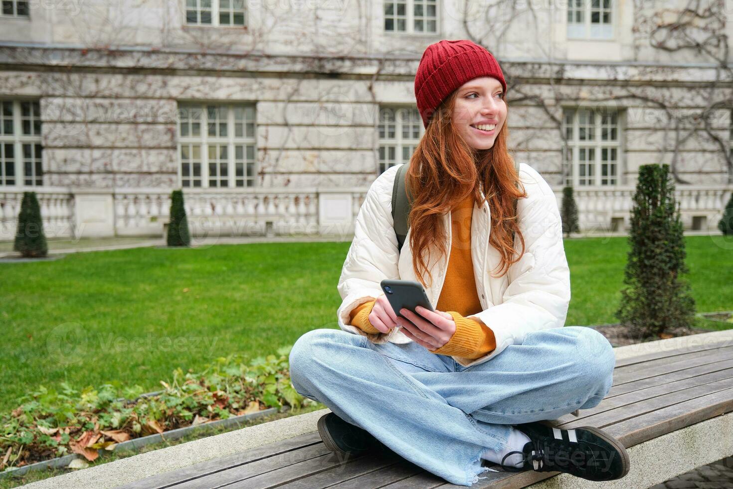 Young smiling redhead girl sits on bench and uses smartphone app, reads news online, watches video on mobile phone while relaxed in park photo
