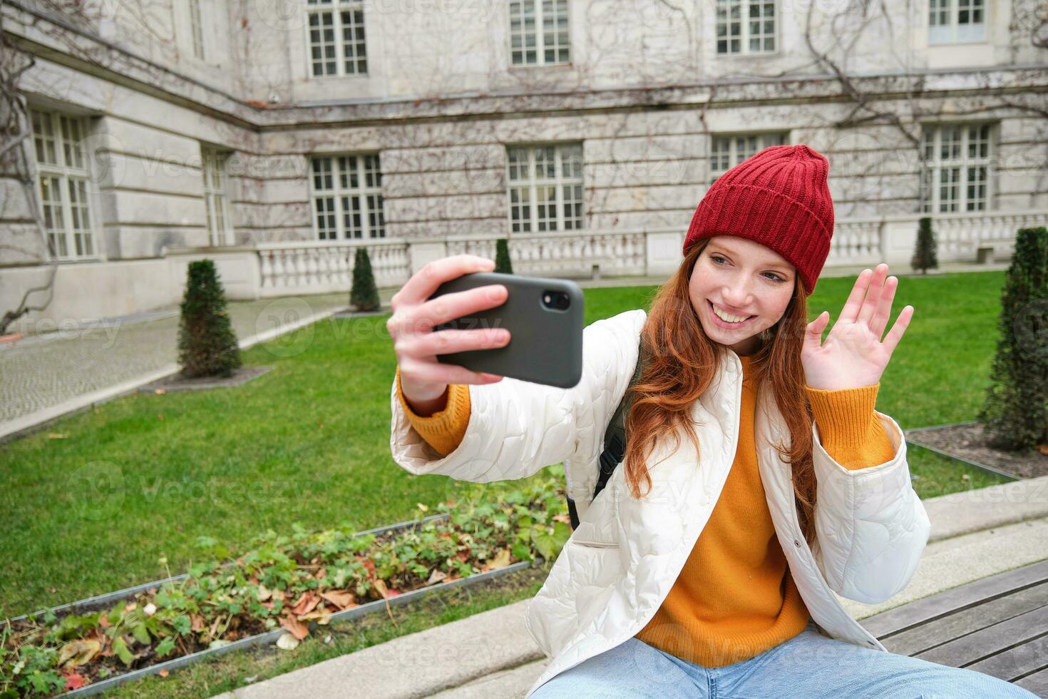 Portrait of girl blogger records video and says hi at phone camera. Young woman takes selfie with smartphone app, waves hand, video chats in application photo