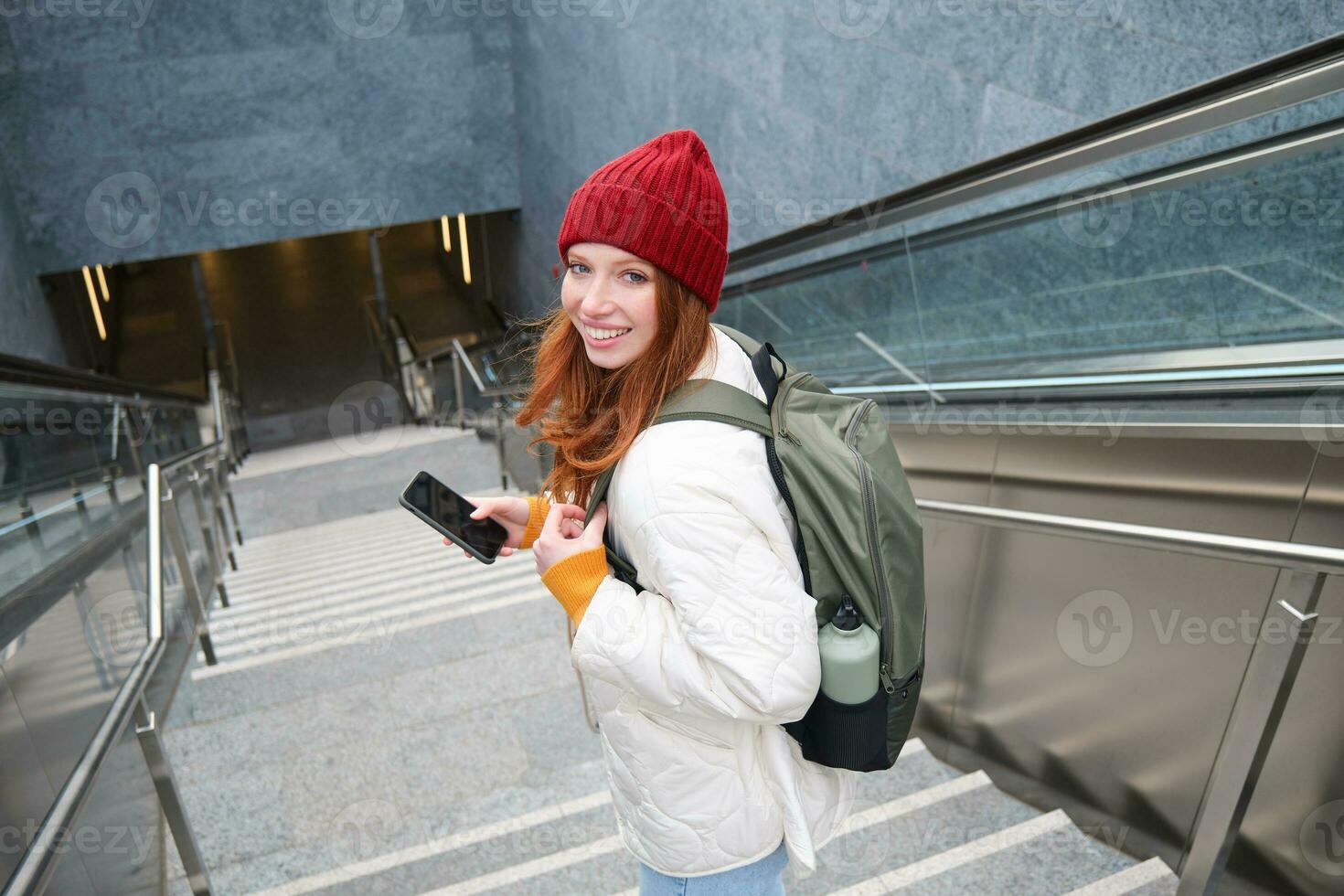 Portrait of redhead girl exploring city with smartphone map app. Young woman using mobile phone and walking around city, going down stairs photo