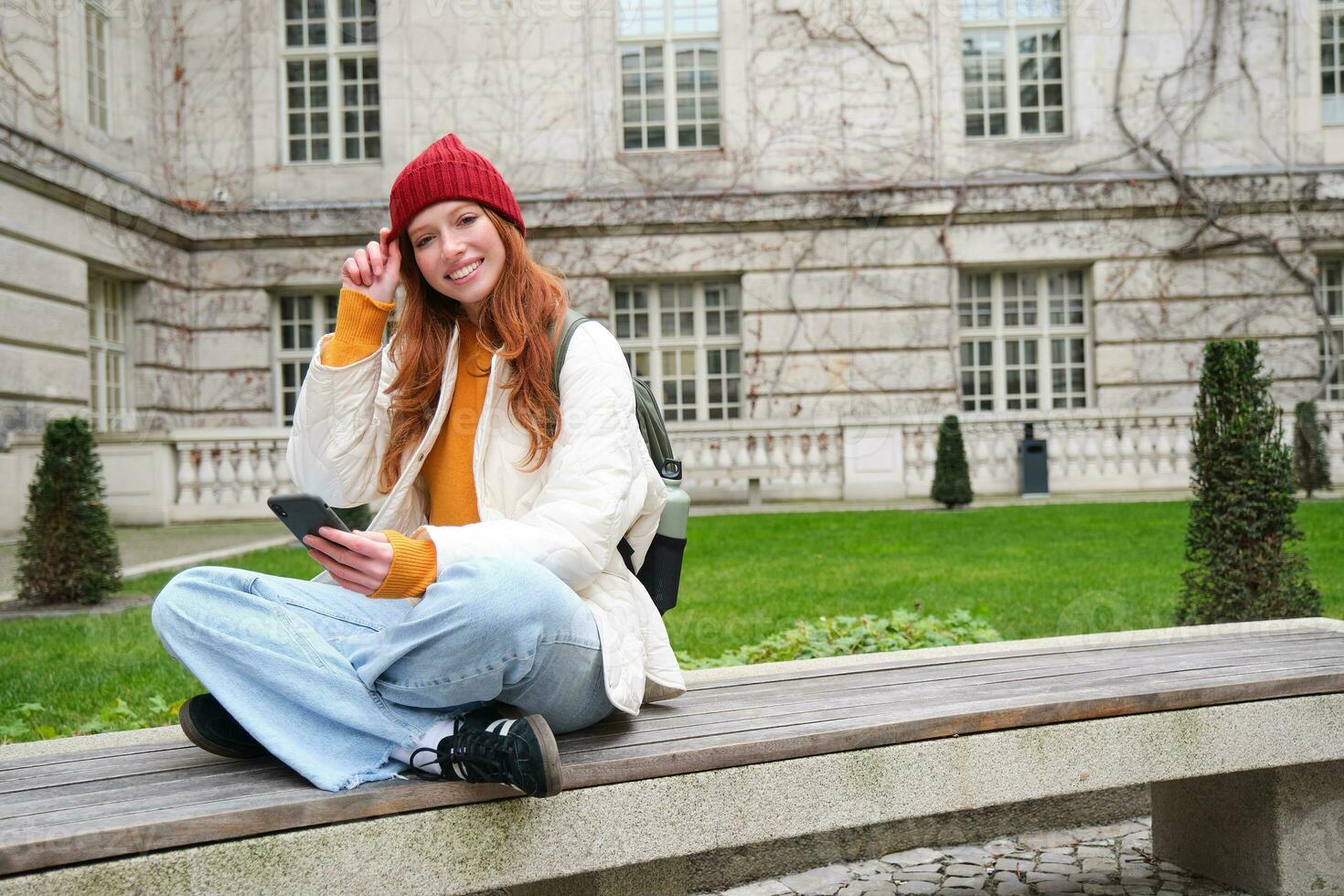 Young smiling redhead girl sits on bench and uses smartphone app, reads news online, watches video on mobile phone while relaxed in park photo