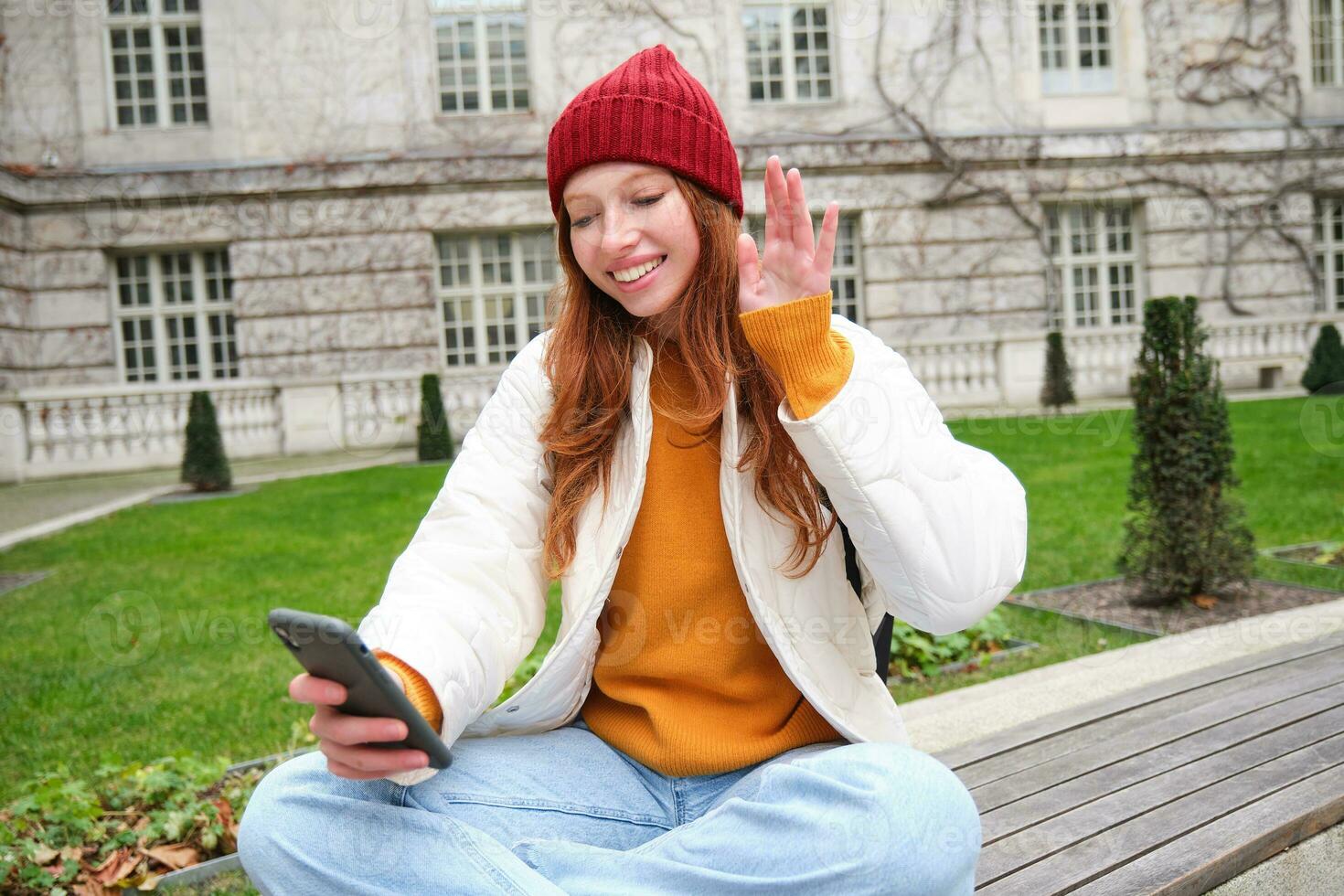 Young smiling redhead girl sits on bench and uses smartphone app, says hi and waves at mobile phone camera, video chats photo