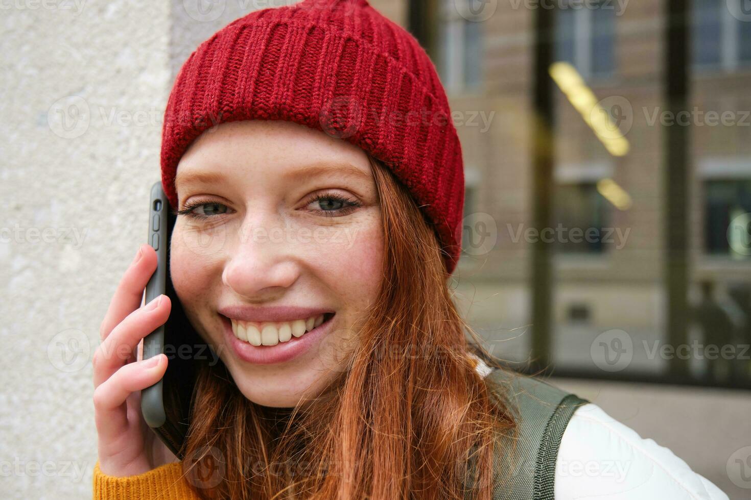 Young people and mobile connection. Happy redhead girl talks on phone, makes telephone call, stands outdoors with backpack and uses smartphone app photo