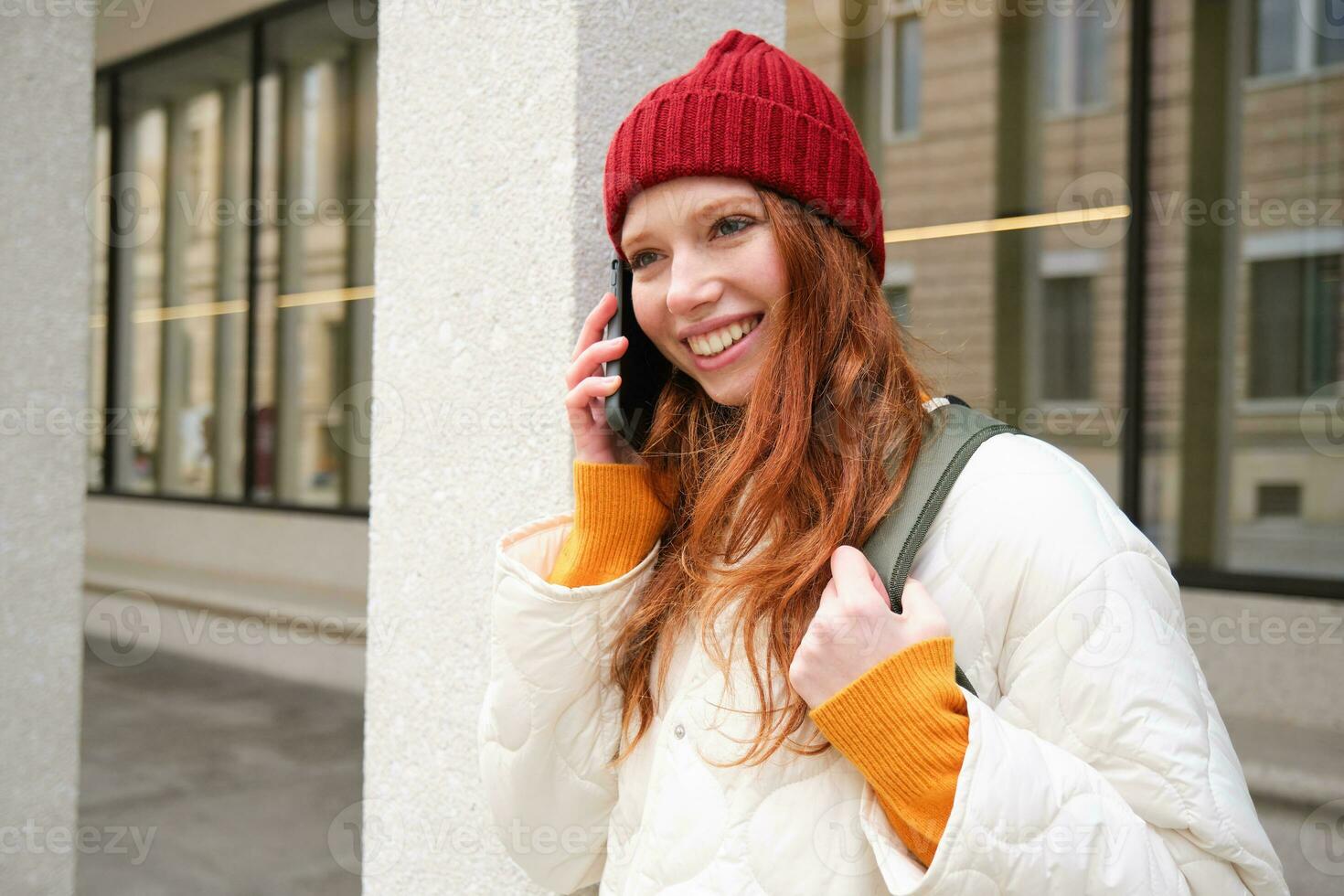Portrait of smiling ginger girl with red hat, stands on street with backpack, rings someone on phone app, talks on mobile, uses smartphone photo