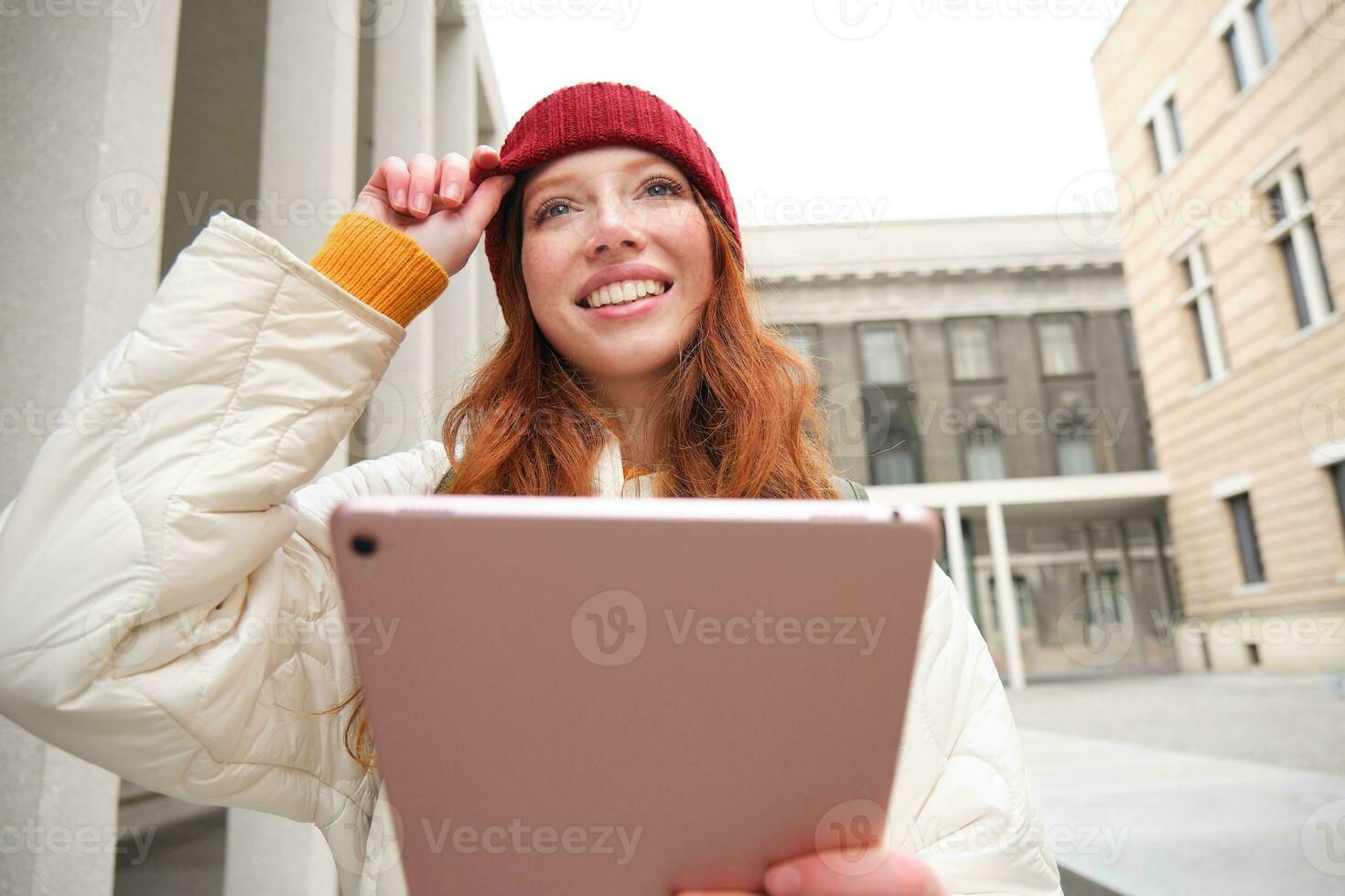 contento pelirrojo niña en rojo sombrero, camina alrededor ciudad con digital tableta, conecta a público Internet Wifi y mira para ruta, mira a mapa en su artilugio foto