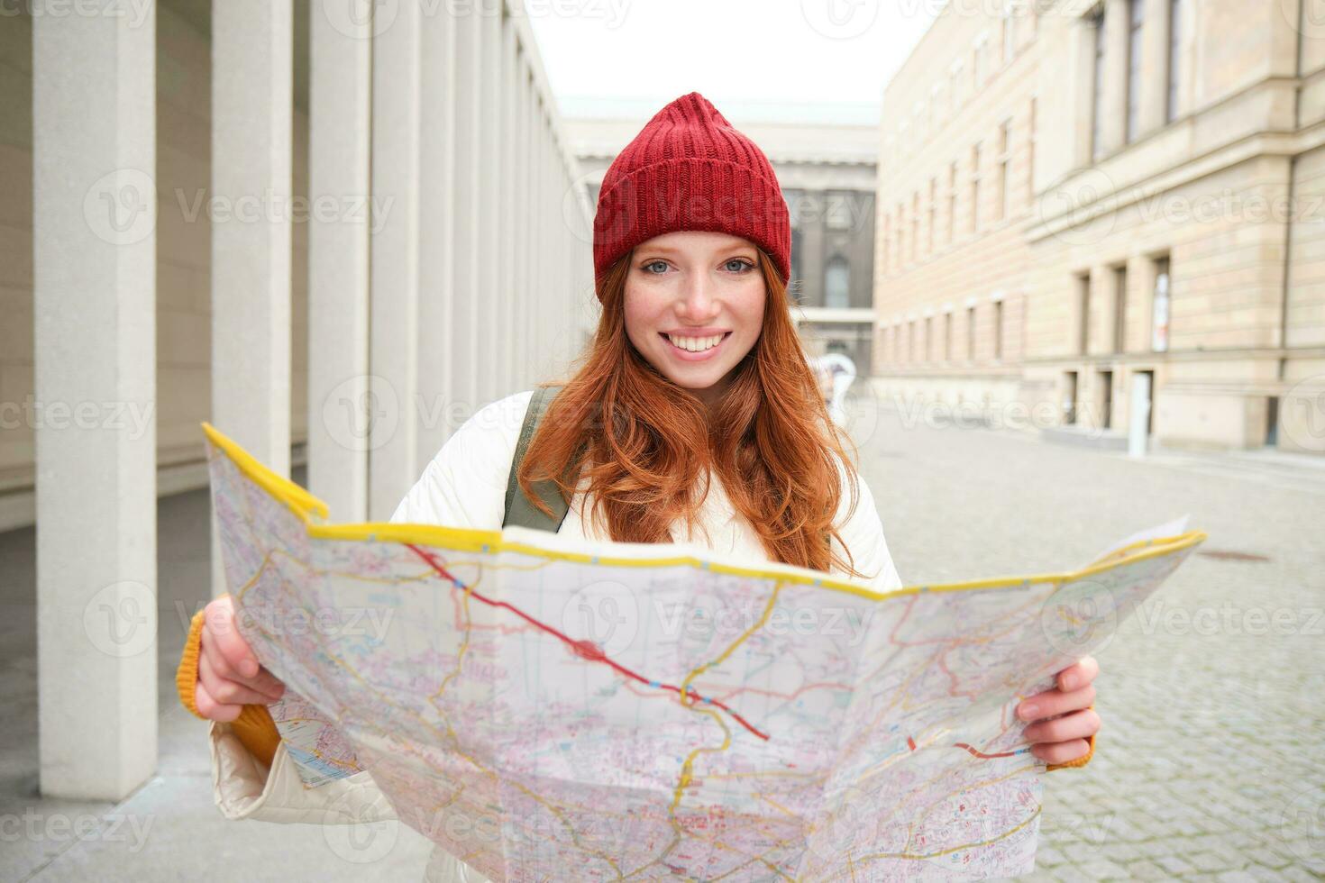 Beautiful redhead woman, tourist with city map, explores sightseeing historical landmark, walking around old town, smiling happily photo