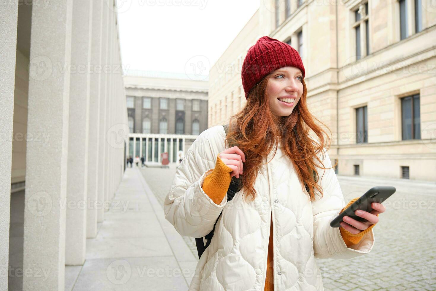 Beautiful smiling girl, tourist with backpack, holding smartphone, using map on mobile phone application, looking for sightseeing in internet app, standing outdoors photo