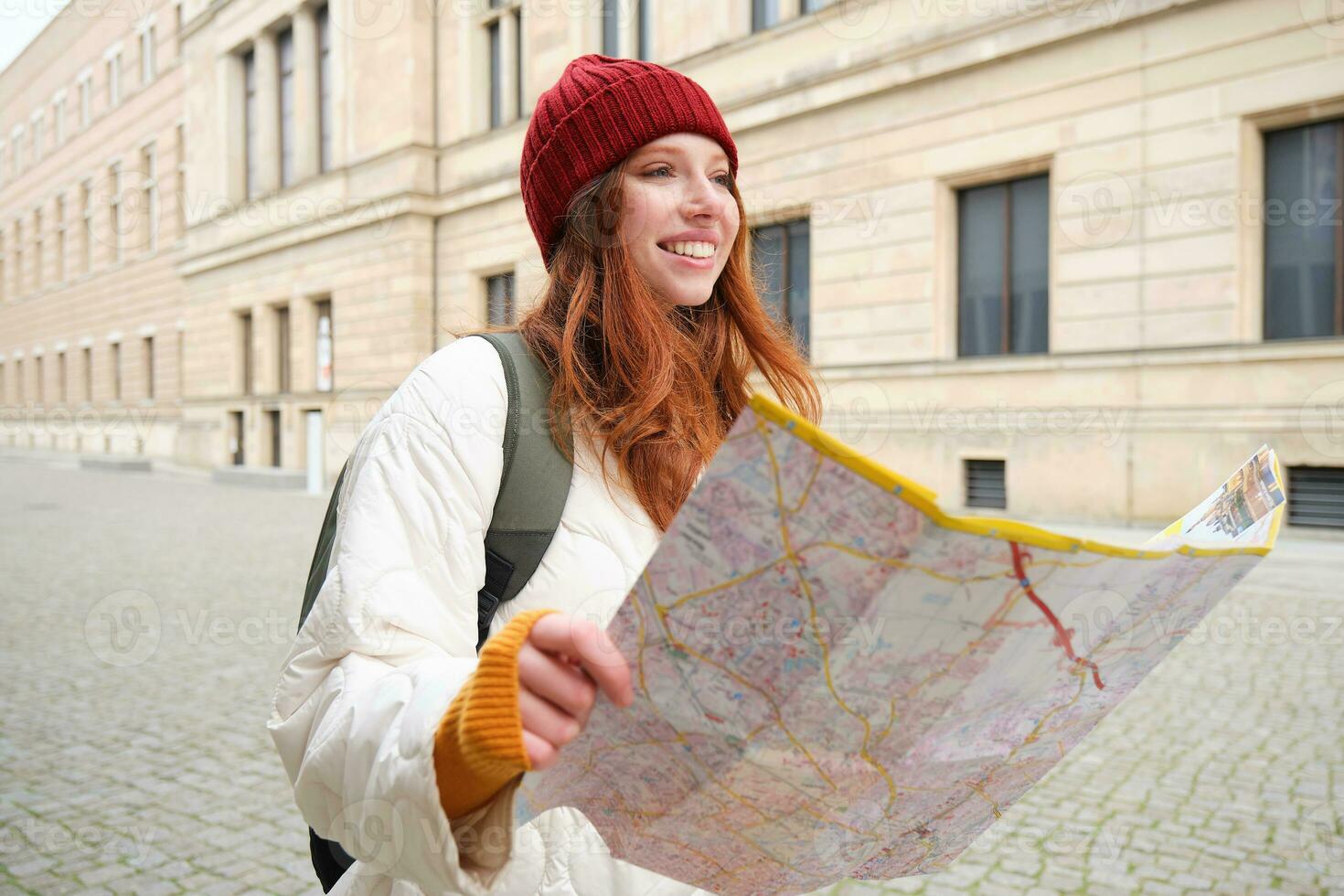 Beautiful redhead woman, tourist with city map, explores sightseeing historical landmark, walking around old town, smiling happily photo