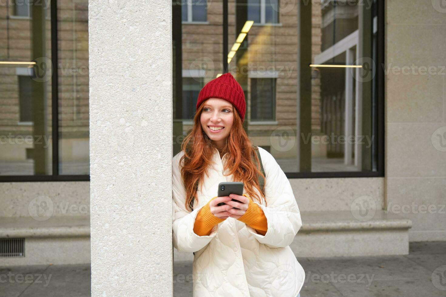 Beautiful redhead girl with smartphone, standing on street, holding mobile phone, using taxi app, connects to public wifi and uses internet, sends a text message photo
