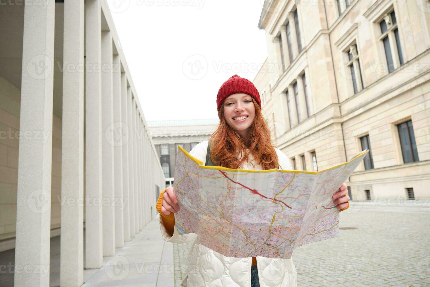 Beautiful redhead woman, tourist with city map, explores sightseeing historical landmark, walking around old town, smiling happily photo