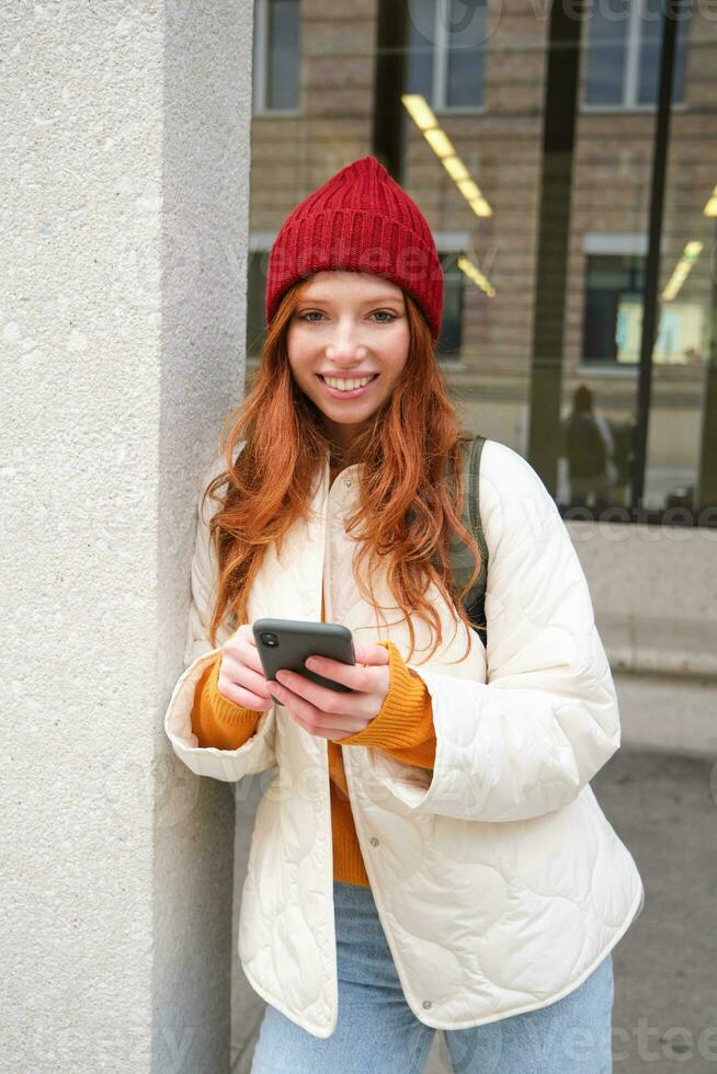 Beautiful redhead girl with smartphone, standing on street, holding mobile phone, using taxi app, connects to public wifi and uses internet, sends a text message photo
