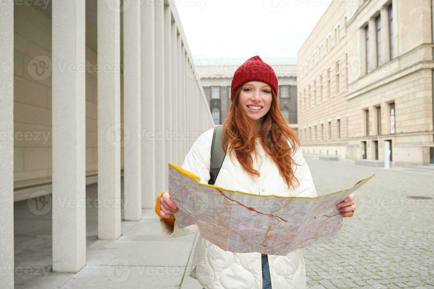 Beautiful redhead woman, tourist with city map, explores sightseeing historical landmark, walking around old town, smiling happily photo