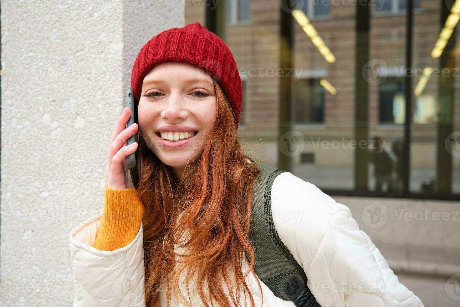 Young people and mobile connection. Happy redhead girl talks on phone, makes telephone call, stands outdoors with backpack and uses smartphone app photo