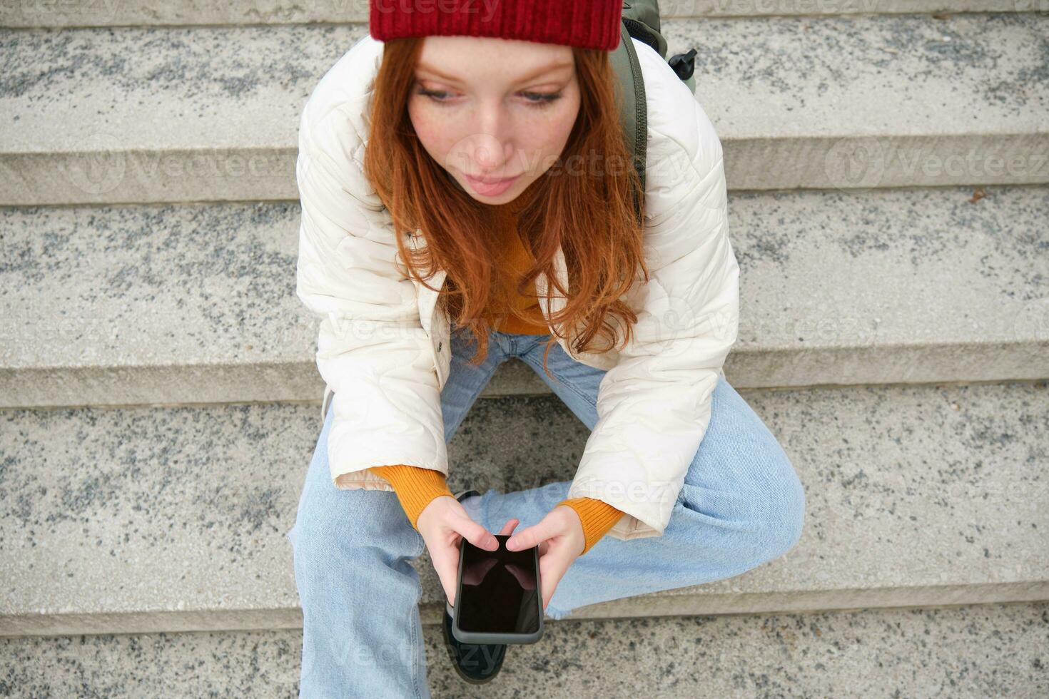 High angle photo of stylish redhead teen girl texts message on phone, uses mobile application while sits outdoors on public stairs