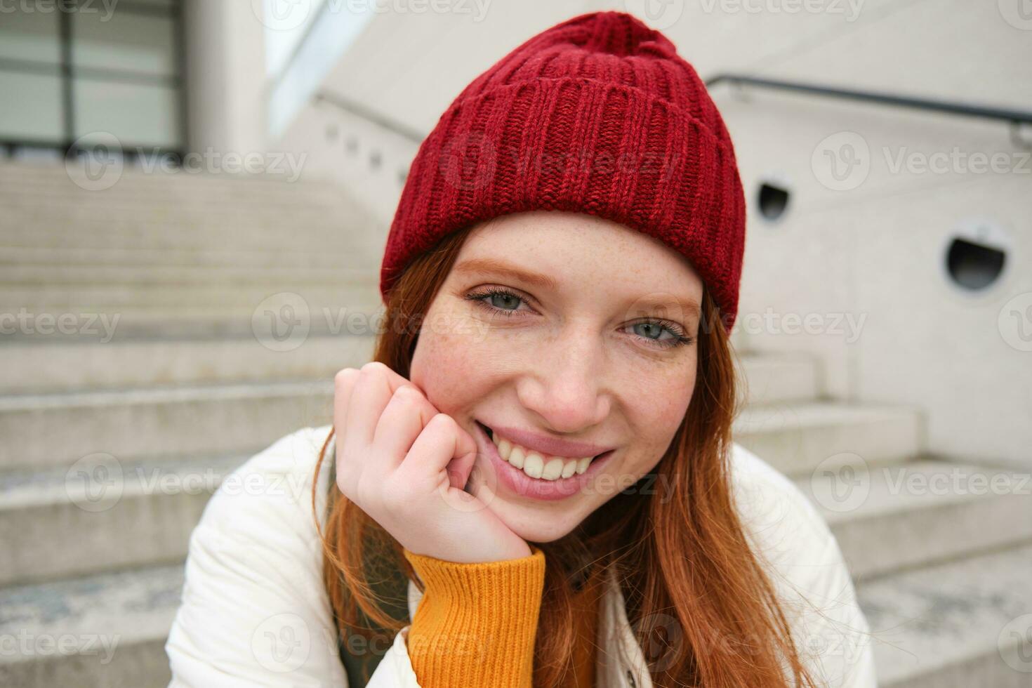 hermosa pelirrojo alumno, niña en rojo sombrero, sonrisas sincero, mira contento y relajado, se sienta en escalera al aire libre foto