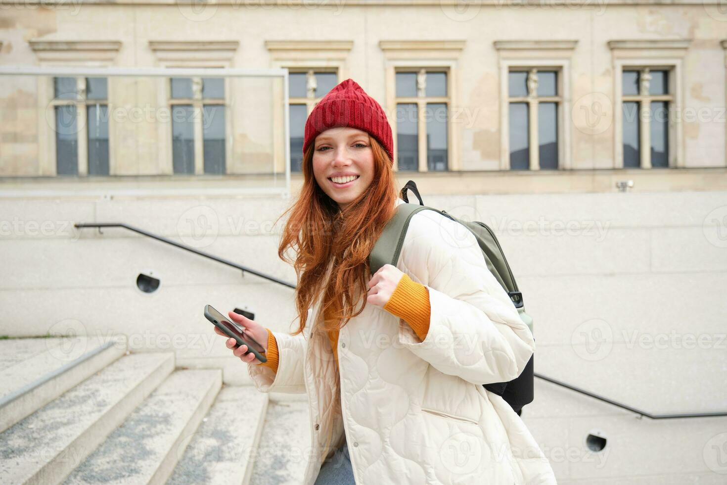 Smiling redhead girl, student tourist walks around city, goes up the stairs, looks at mobile phone map to get around town, sends message on smartphone photo