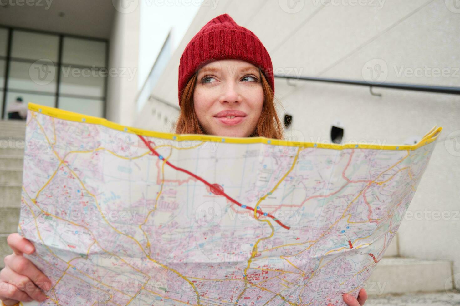 Young smiling redhead girl, tourist sits on stairs outdoors with city paper map, looking for direction, traveller backpacker explores city and looks for sightseeing photo