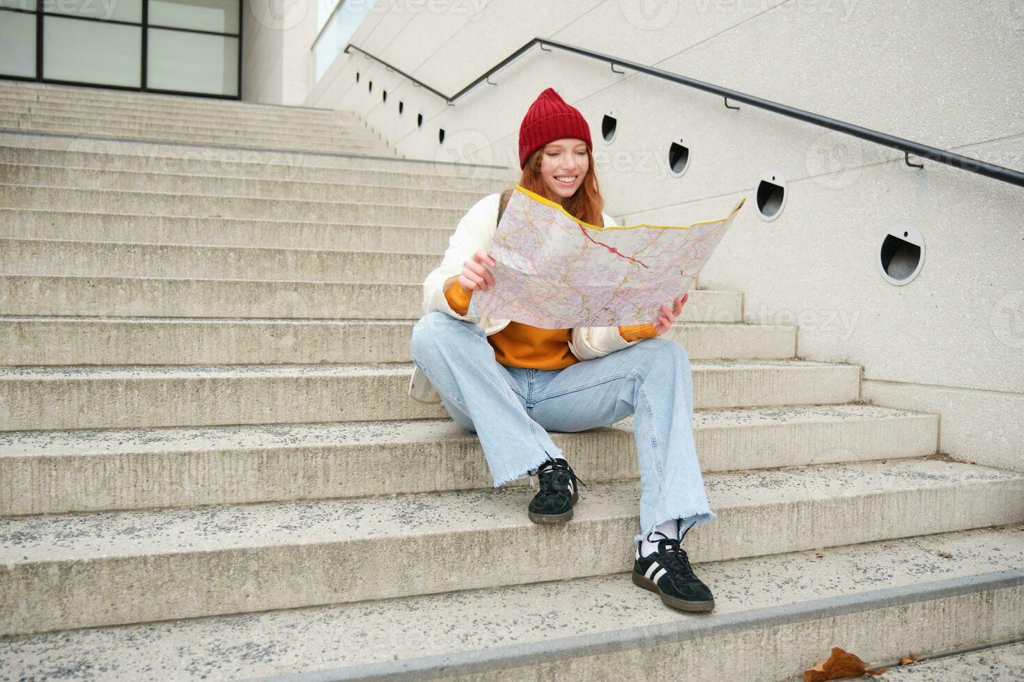 Young smiling redhead girl, tourist sits on stairs outdoors with city paper map, looking for direction, traveller backpacker explores city and looks for sightseeing photo