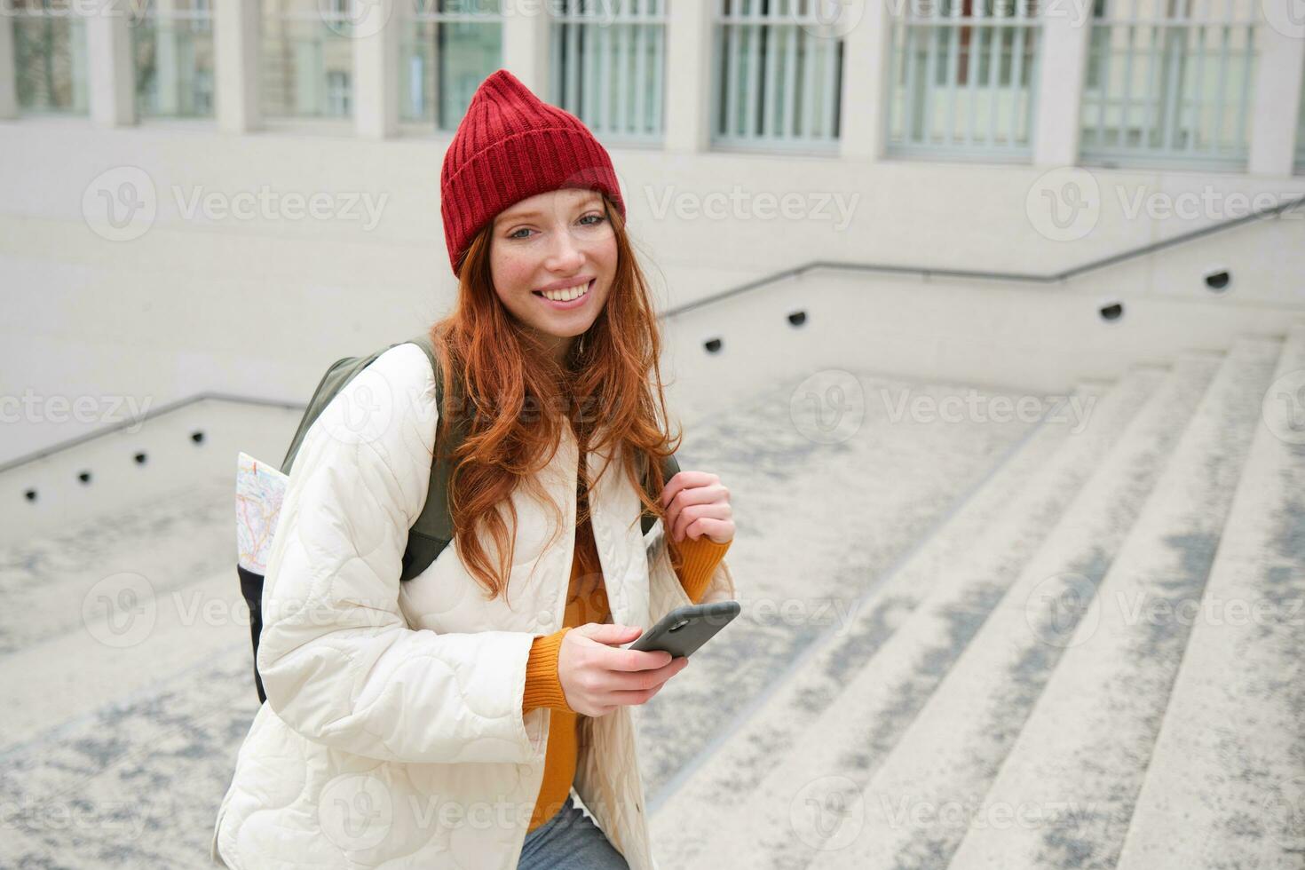 Smiling redhead girl, student tourist walks around city, goes up the stairs, looks at mobile phone map to get around town, sends message on smartphone photo