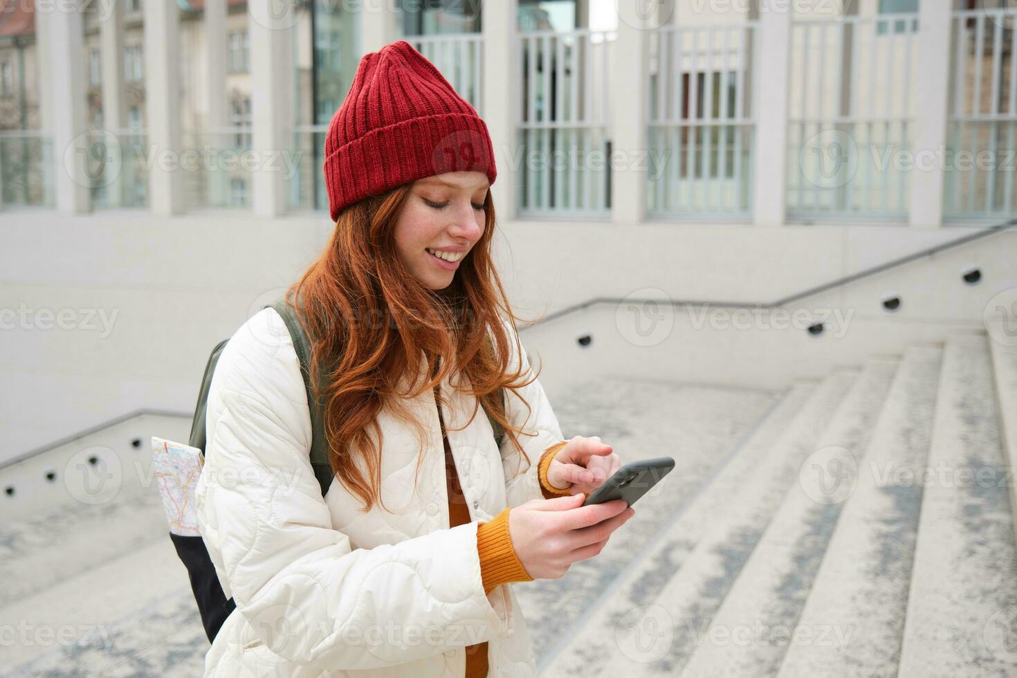 Smiling redhead girl, student tourist walks around city, goes up the stairs, looks at mobile phone map to get around town, sends message on smartphone photo