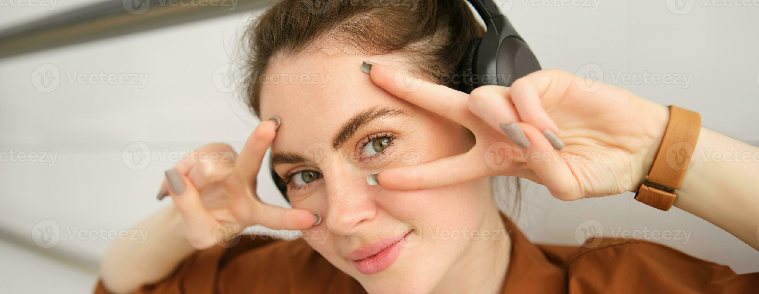 Close up portrait of woman in headphones, shows peace, vsigns near eyes, smiles and looks happy, sits on floor at home photo