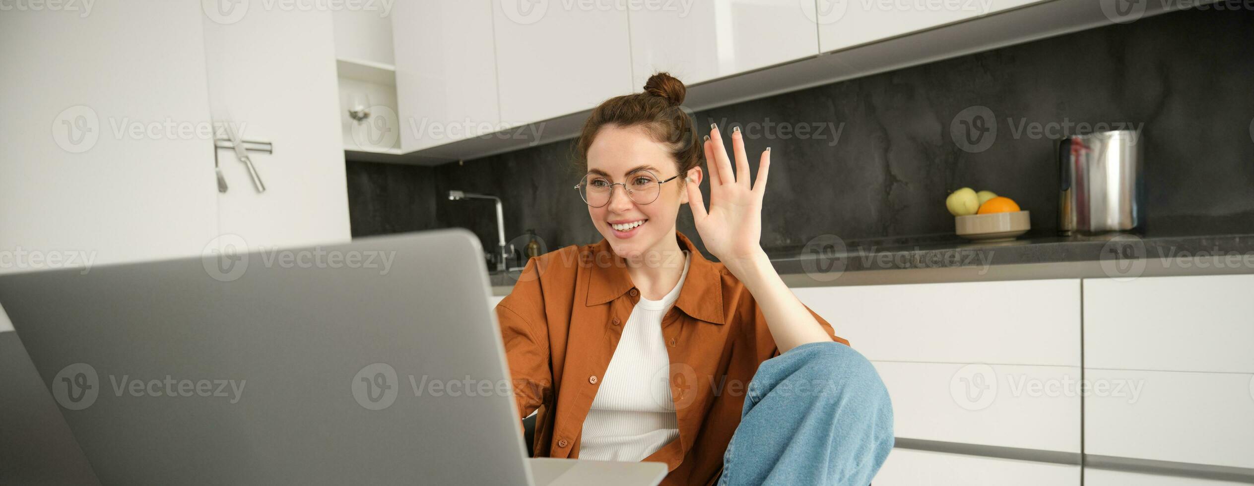 alegre joven mujer en anteojos, diciendo Hola, ondulación mano a computadora portátil, hablando a amigo en línea, vídeo charlando, sentado en cocina con computadora foto