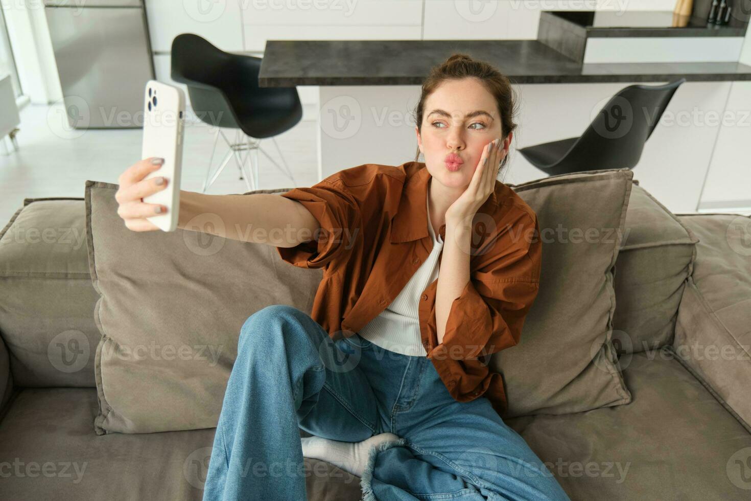 Portrait of cute smiling female model, takes selfie on smartphone app, posting photos on social media, using mobile phone camera, posing on sofa at home
