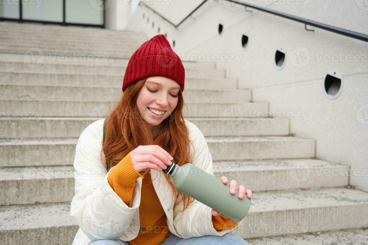 Smiling traveler, redhead girl tourist sits on stairs with flask, drinks hot coffee from thermos while travelling and sightseeing around foreign city, sits on stairs and rests photo