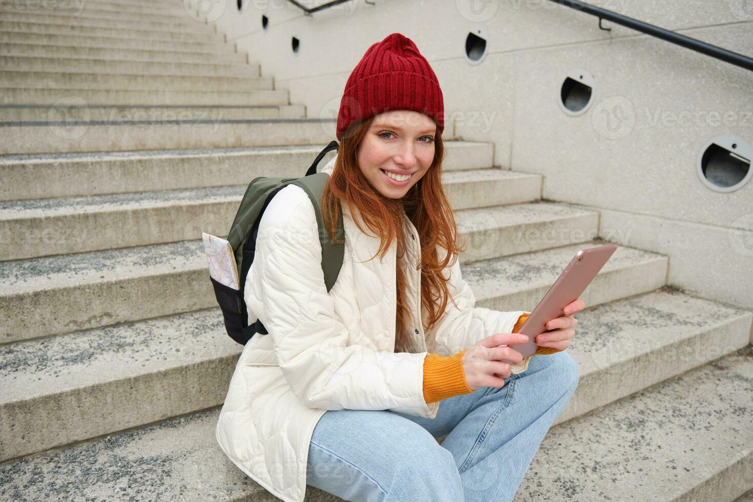 Happy stylish redhead girl, student in red hat, holds digital tablet, uses social media app, searches something online, connects to wifi photo