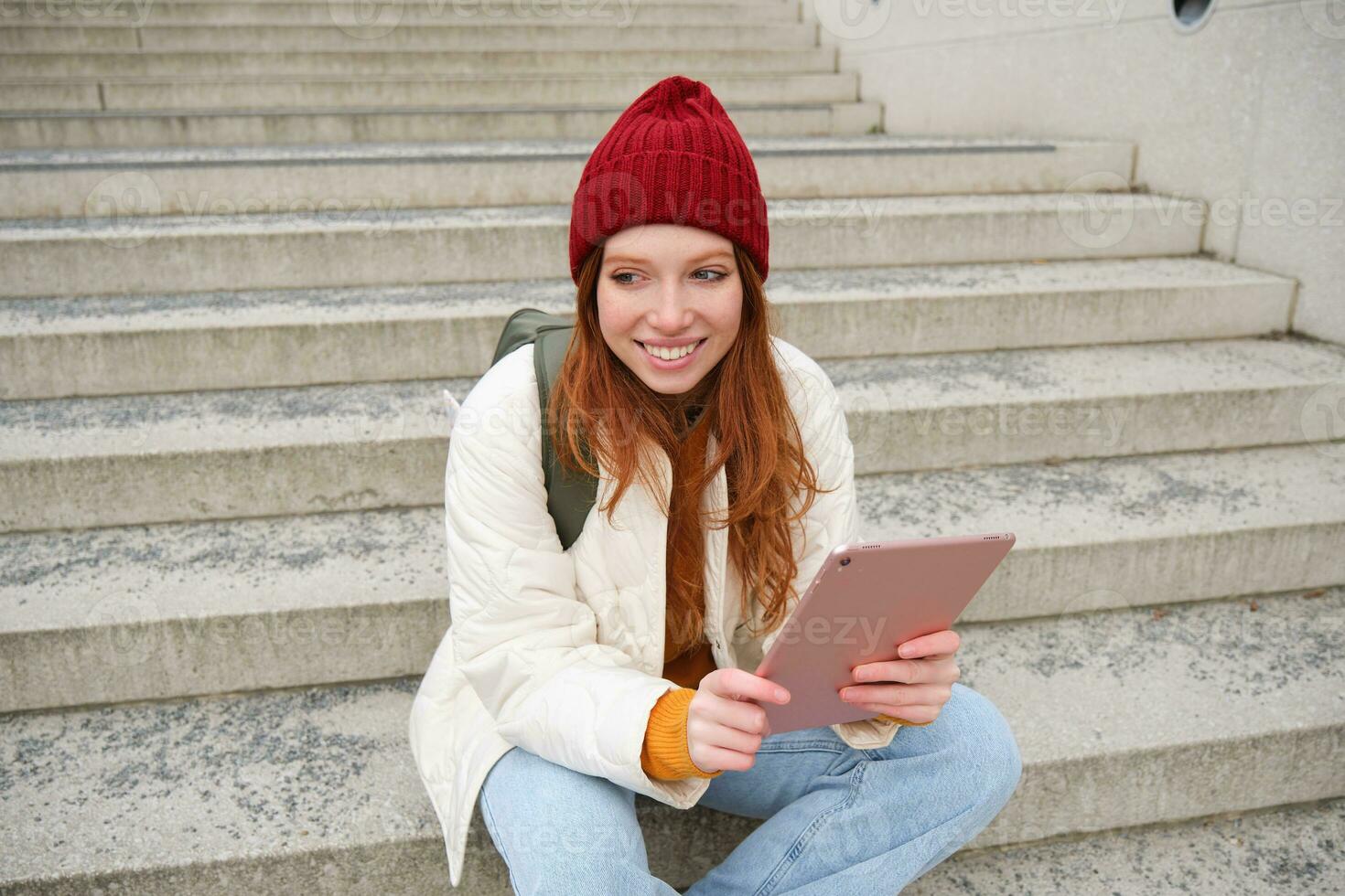 Young stylish girl, redhead female students sits on stairs outdoors with digital tablet, reads, uses social media app on gadget, plays games while waits on street photo