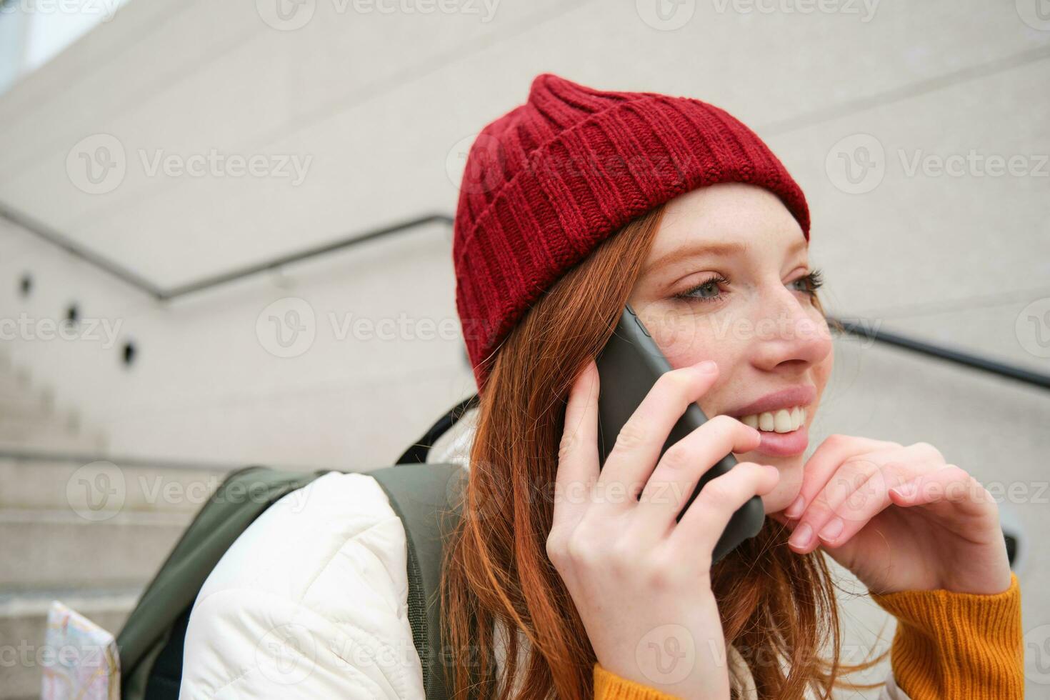 hermosa sonriente pelirrojo hembra modelo, se sienta en calle y negociaciones en móvil teléfono, usos teléfono inteligente aplicación a llamada en el extranjero, riendo durante teléfono conversacion foto