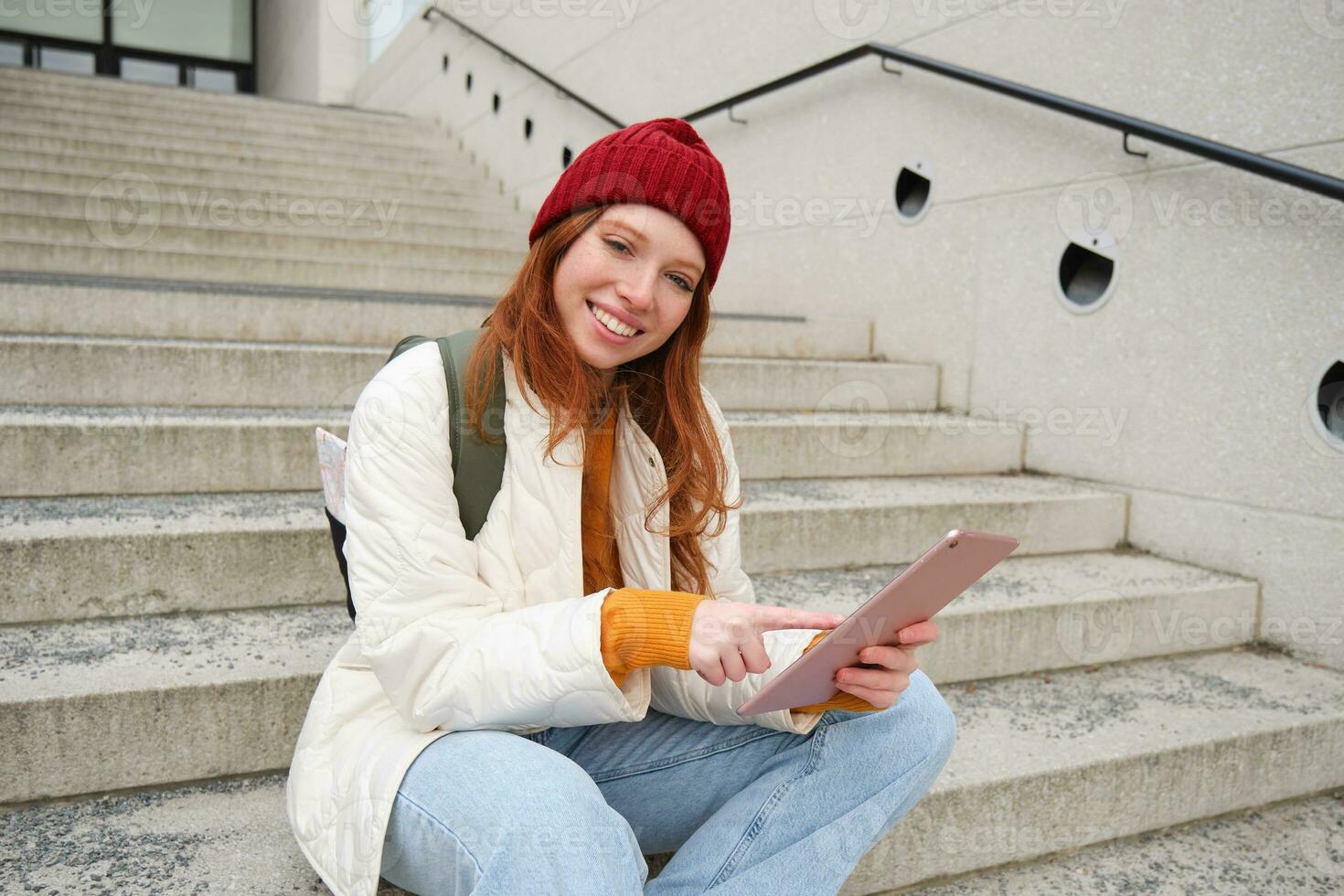Happy stylish redhead girl, student in red hat, holds digital tablet, uses social media app, searches something online, connects to wifi photo