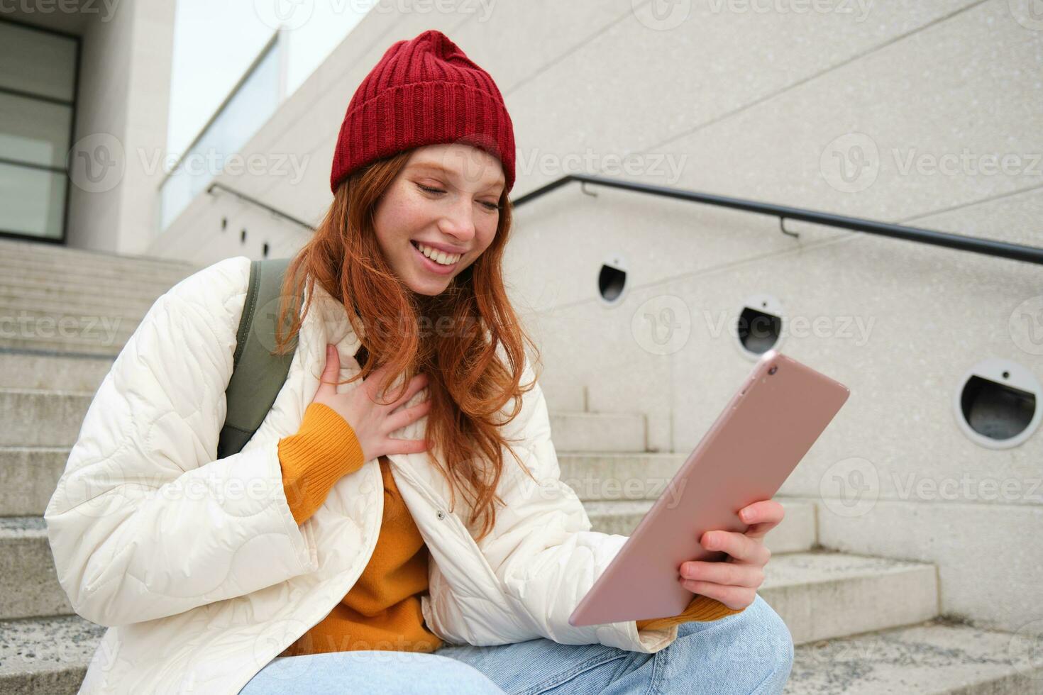 Happy stylish redhead girl, student in red hat, holds digital tablet, uses social media app, searches something online, connects to wifi photo