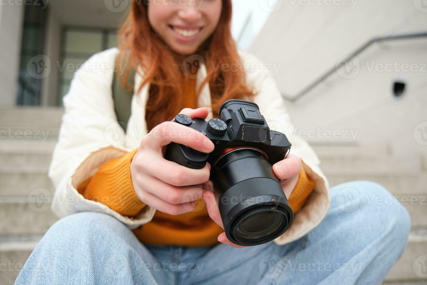 sonriente pelirrojo niña fotógrafo, cheques su tiros, sostiene cámara y mira a pantalla, toma fotos al aire libre, camina alrededor calle y lo hace estilo callejero disparo