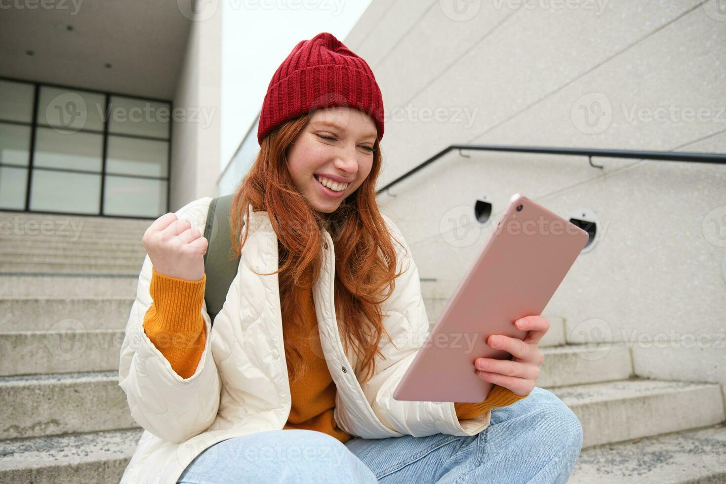 Young stylish girl, redhead female students sits on stairs outdoors with digital tablet, reads, uses social media app on gadget, plays games while waits on street photo