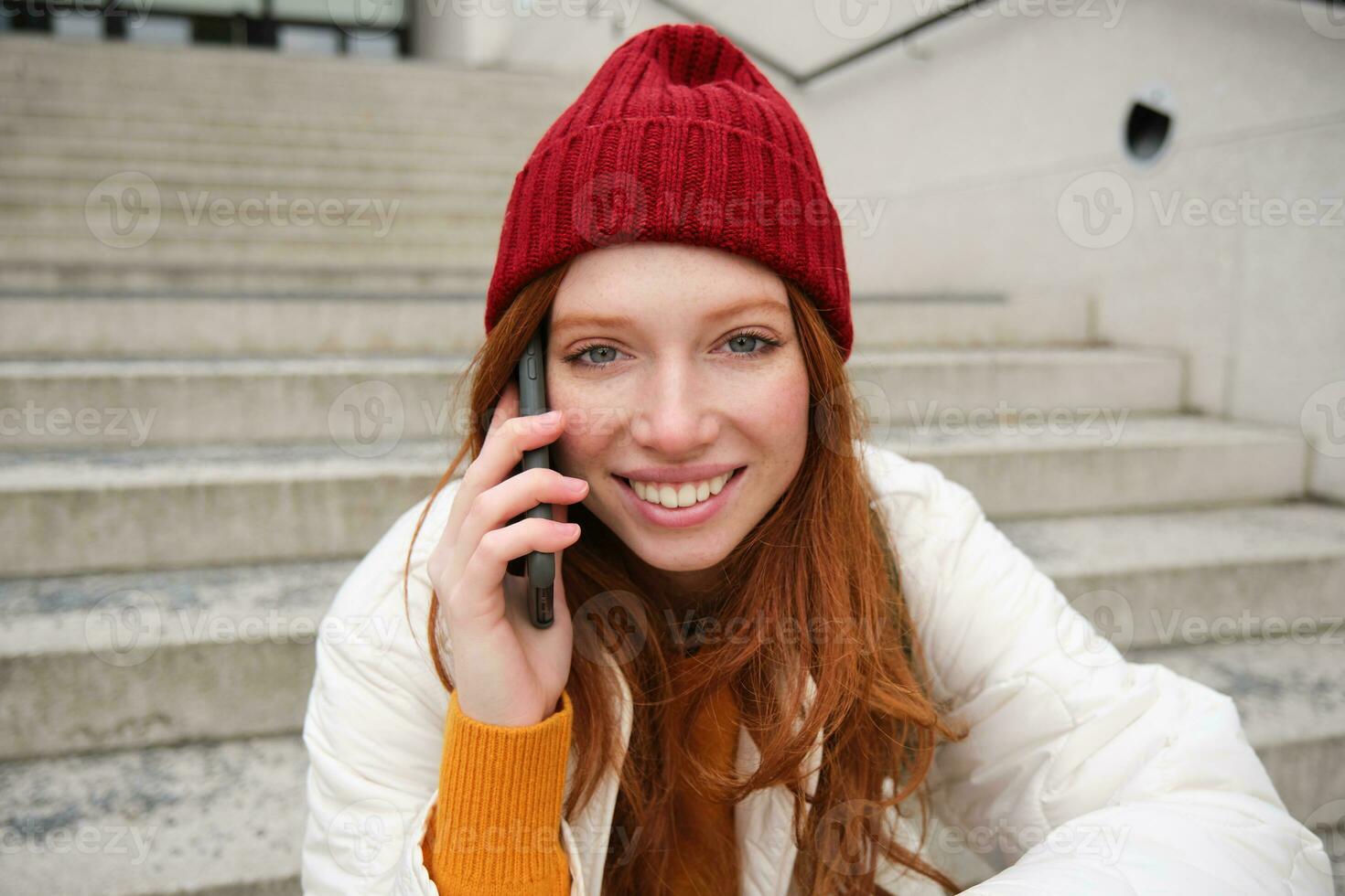 hermosa sonriente pelirrojo hembra modelo, se sienta en calle y negociaciones en móvil teléfono, usos teléfono inteligente aplicación a llamada en el extranjero, riendo durante teléfono conversacion foto