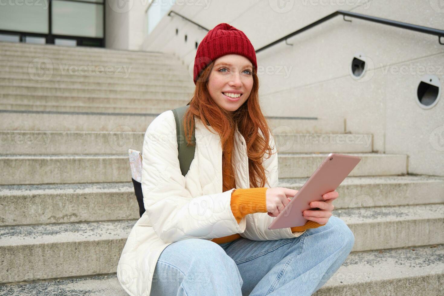 Young stylish girl, redhead female students sits on stairs outdoors with digital tablet, reads, uses social media app on gadget, plays games while waits on street photo