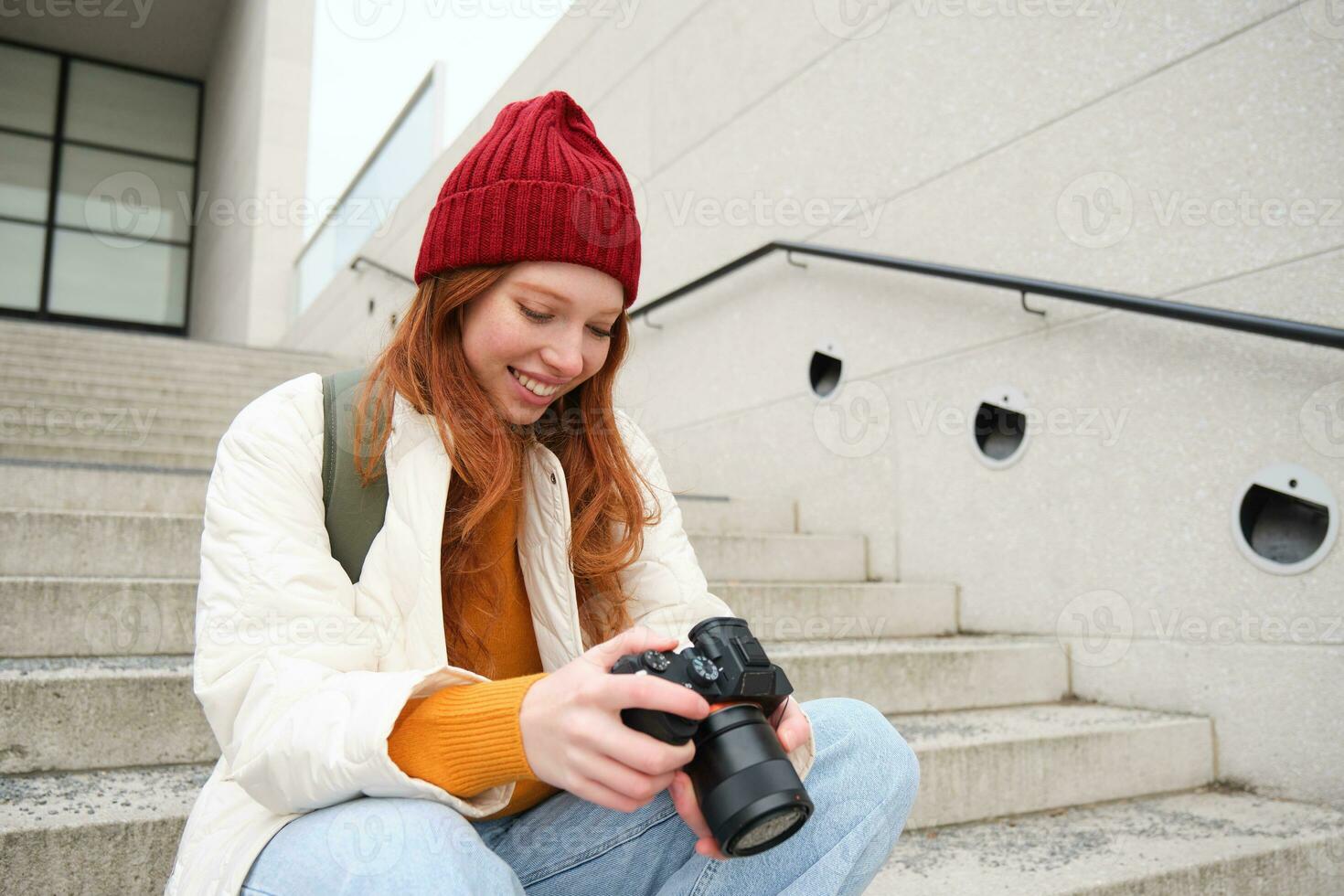 Smiling redhead girl photographer, checks her shots, holds camera and looks at screen, takes photos outdoors, walks around street and does streetstyle shooting