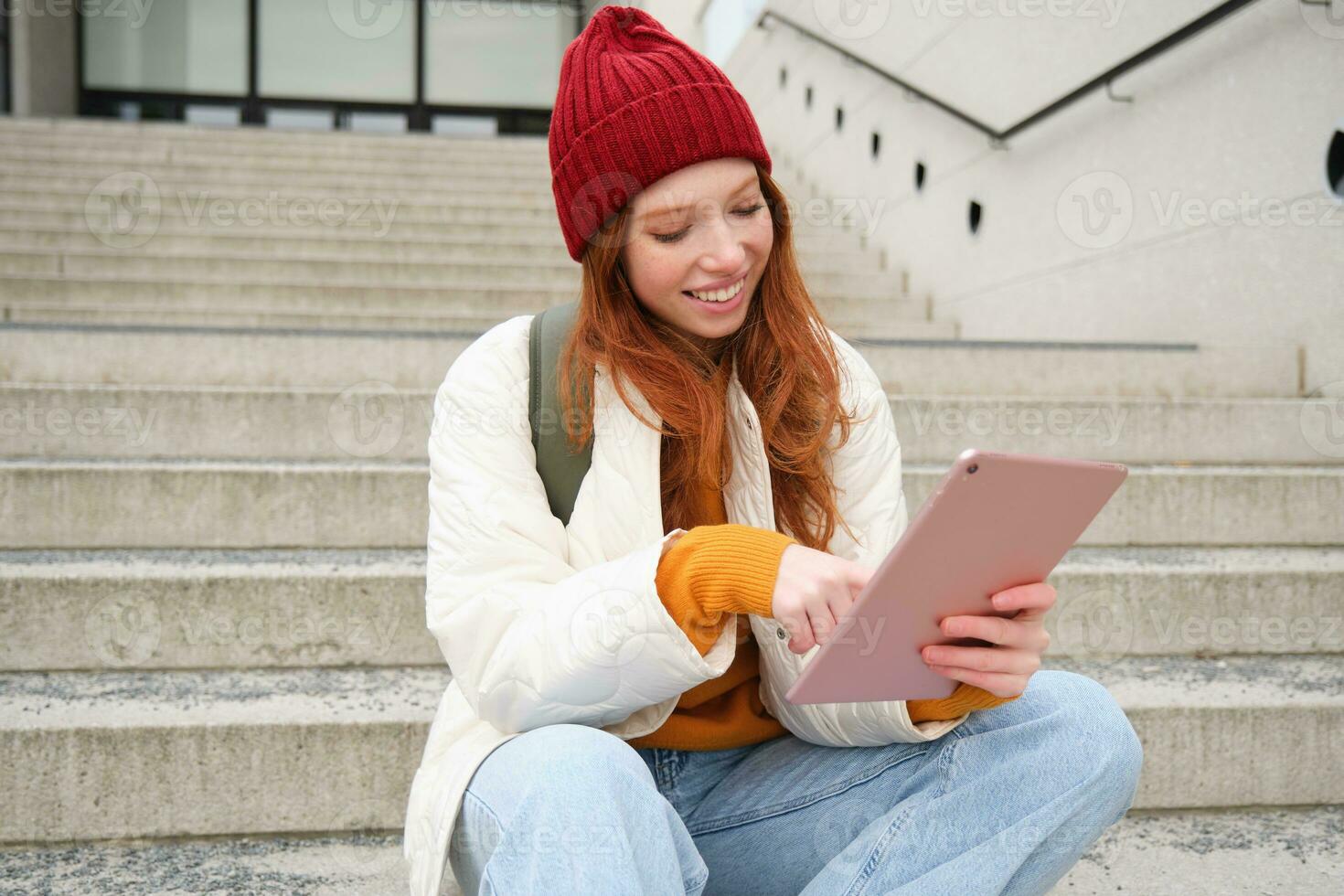 Happy stylish redhead girl, student in red hat, holds digital tablet, uses social media app, searches something online, connects to wifi photo