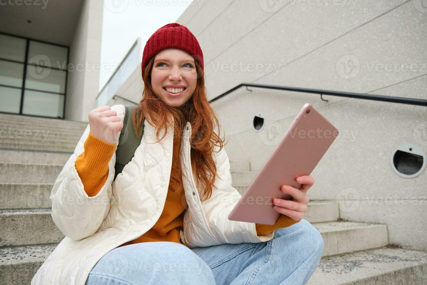 Happy stylish redhead girl, student in red hat, holds digital tablet, uses social media app, searches something online, connects to wifi photo